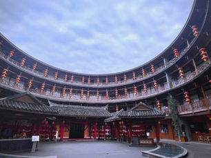Circular courtyard with red lanterns and traditional roofs, under a cloudy sky. Wooden balconies decorated with lanterns encircle the area.