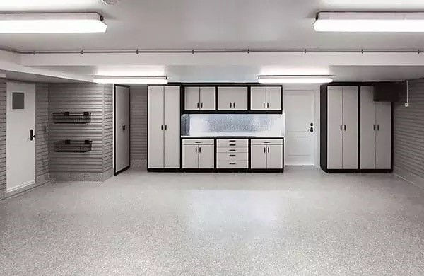 Organized garage with gray cabinets, shelves, and a white door. Bright overhead lights illuminate the clean, spacious, gray-floored area.