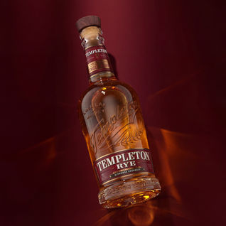Tilted bottle of Templeton Rye whiskey illuminated by a strong spotlight, casting intricate amber reflections and light caustics against a dark red background.