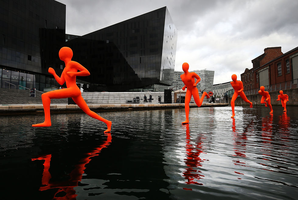 Bright orange sculptures of running figures appear to sprint across the water of a dock on Liverpool's waterfront, with the angular black Mann Island buildings behind them.