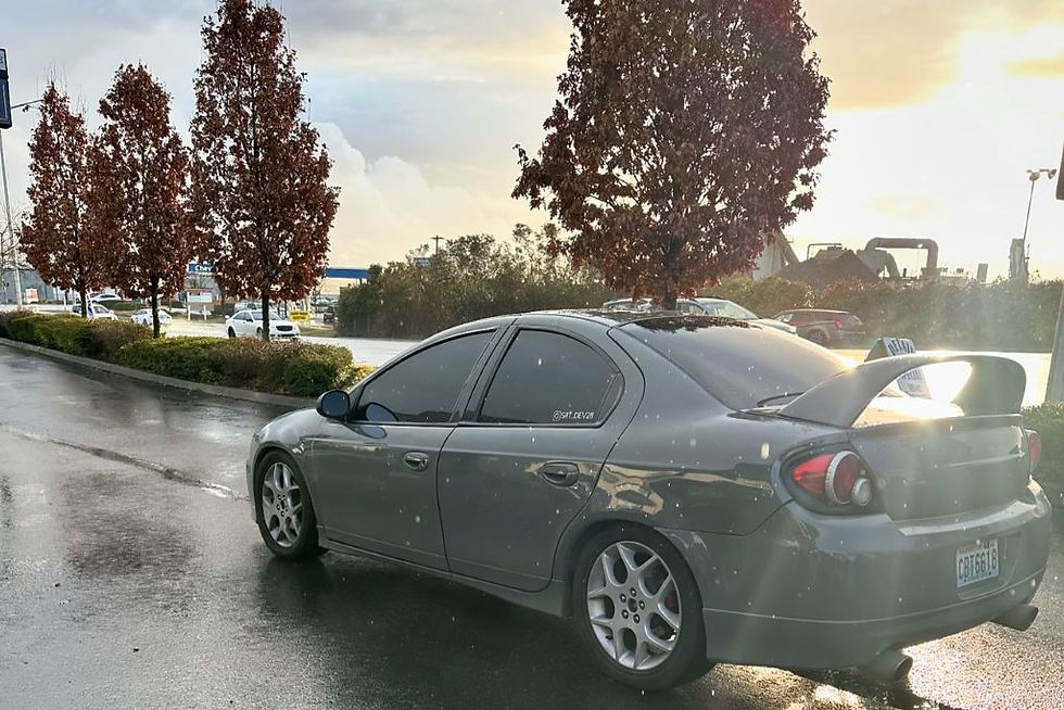 A dark gray sedan with a rear spoiler parked on a wet Tacoma street after rainfall, reflecting overcast light and surrounded by autumn trees. This image illustrates why drivers in Tacoma might ask, "Can PPF cover car scratches?"—due to constant exposure to moisture, debris, and urban wear.