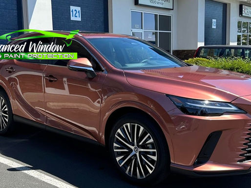 A shiny, rose gold Lexus SUV parked in front of a detailing shop, with dark tinted windows reflecting the sun. The shop has signage reading "Advanced Window Tint & Paint Protection" with a bright green graphic of a car above the text, emphasizing automotive window tinting services.