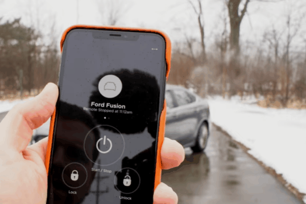 A driver holds a phone showing remote start controls for a Ford Fusion while the car sits on a snowy road. The screen includes "Ford Fusion" "Remote Stopped at 11.22am" "Start. Stop" "Lock" and "Unlock."
