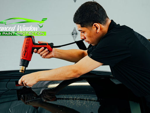 A technician applies window tint to a car using a heat gun and squeegee, focusing carefully on the rear window to smooth the film. The image includes the logo for "Advanced Window Tint & Paint Protection" in green and white.