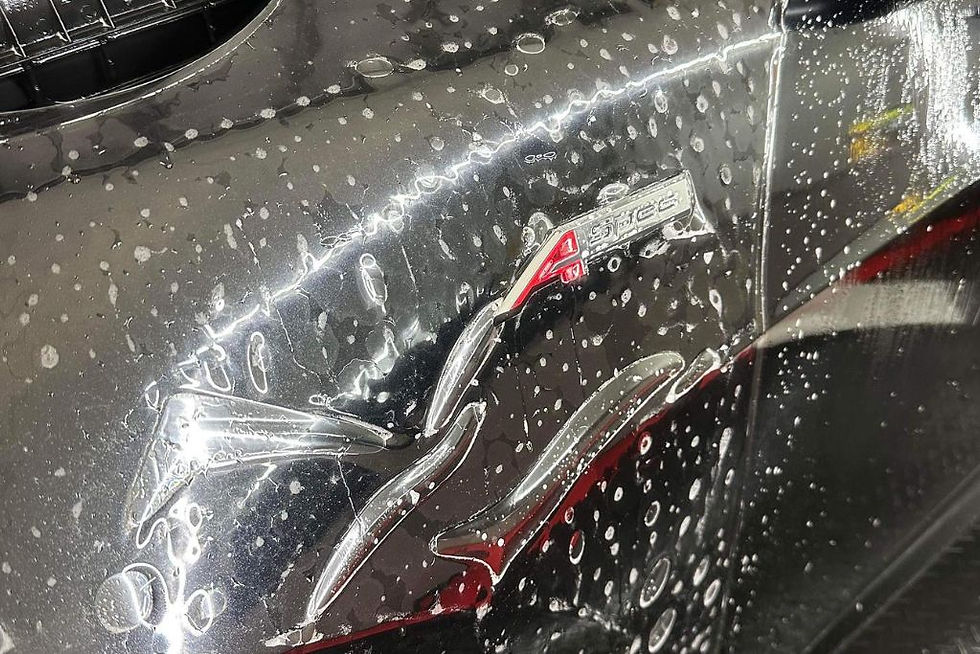 Close-up of a black car panel with water droplets and partially applied paint protection film (PPF) over the “A-Spec” badge. The clear film highlights how PPF conceals minor scratches and swirl marks during the installation process.