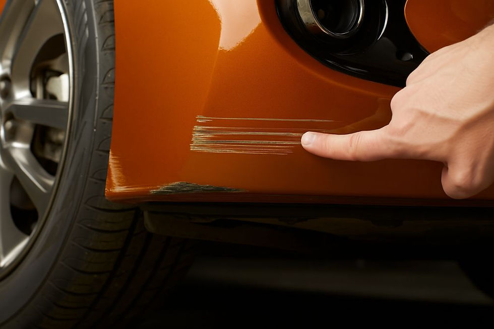 A person points to multiple scratches and scuffs on the lower bumper of an orange car, emphasizing areas where Paint Protection Film would not be effective in concealing deep paint damage.