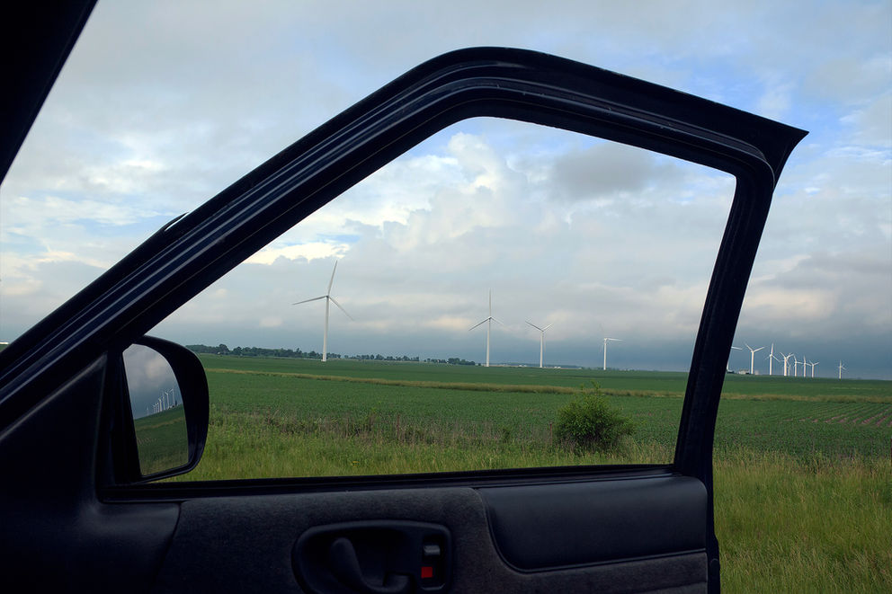 A scenic roadside view of a Midwestern wind farm, seen through the open door of a parked vehicle. Wind turbines stand tall over lush green farmland, under a cloudy sky. The location is likely Illinois or a neighboring state, known for its extensive wind energy infrastructure and rural highways.