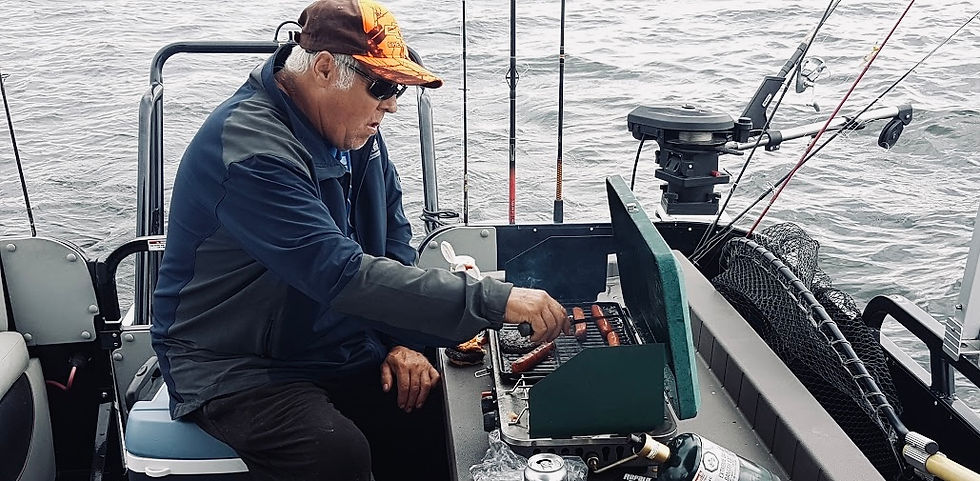 A man wearing a baseball cap, sunglasses, and a dark jacket grills sausages on a portable barbecue aboard a fishing boat. Several fishing rods and a landing net are visible, with the boat surrounded by open water.