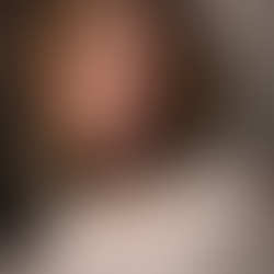 Smiling woman, Sheridan Guerrette, with wavy hair in white lace top, wearing a long necklace and scarf, stands against a tiled wall. Warm and relaxed mood.