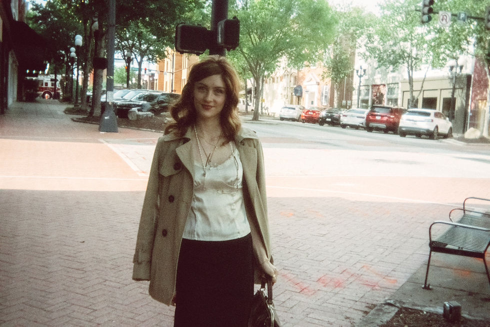 A woman, Sheridan Guerrette, in a coat stands on a sunny city street, smiling. Background shows trees, parked cars, and brick buildings. A calm, pleasant scene.