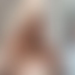 A woman, Sheridan Guerrette, with a towel wrapped on her head and red nails poses in a bathroom with white and black tiled walls, looking directly at the camera.