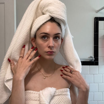 A woman, Sheridan Guerrette, with a towel wrapped on her head and red nails poses in a bathroom with white and black tiled walls, looking directly at the camera.