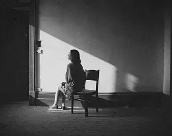 Black-and-white photo of a woman sitting alone on a chair in an empty room, lit by a sharp sunbeam on the wall.