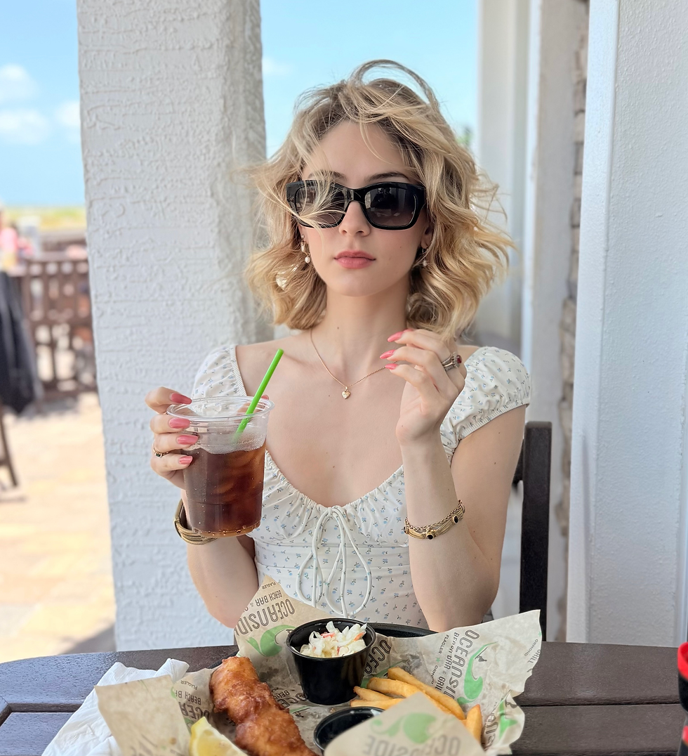 Woman, aka sheridan guerrette, with sunglasses sips a drink at an outdoor table. Fish, fries, and coleslaw on plate. Sunny day; casual, relaxed mood.