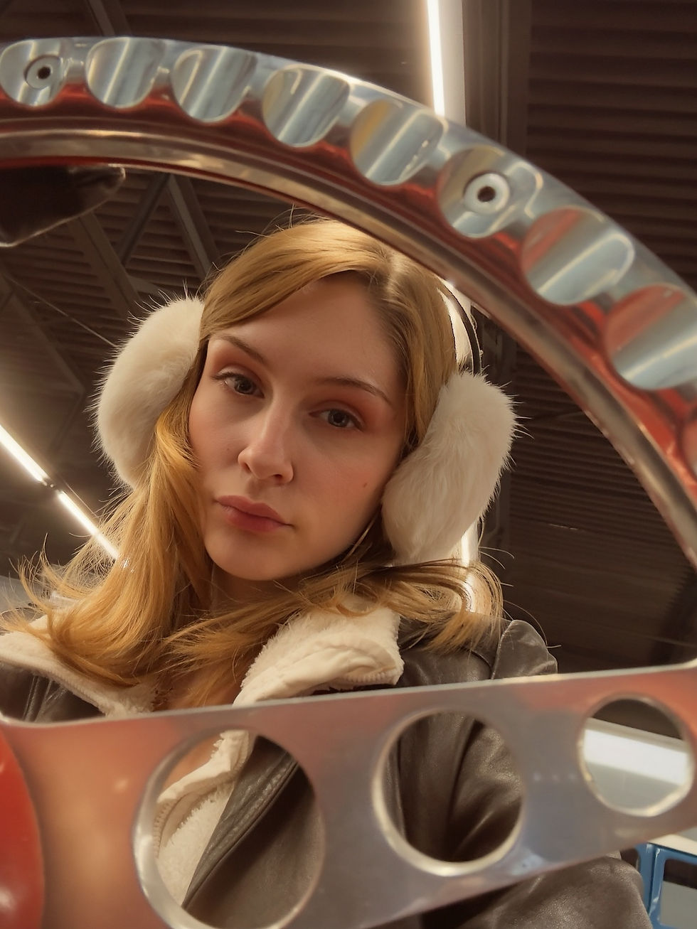 Sheridan Guerrette wearing white earmuffs and a leather jacket looks through a metal object with circular holes, set against a dark industrial ceiling.