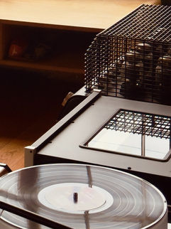 A vinyl record on a turntable spins next to audio equipment in a warmly lit room with wooden flooring. Photograph by American artist and writer Sheridan Guerrette