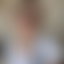 Sheridan Guerrette with sunglasses and white blouse poses in front of a textured concrete wall. She's wearing earrings and necklaces, with a striped bag.