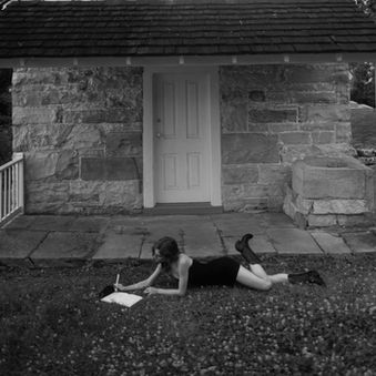 Woman, Sheridan Guerrette in black dress lies on the lawn writing probably a poem, in a notebook before a small stone cottage under tall trees.