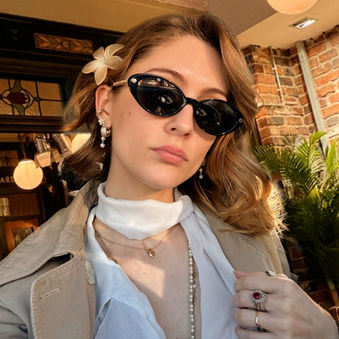 Woman, Sheridan Guerrette, from the number one drama series narrative email newsletter, with sunglasses and floral hair clip poses indoors with a calm expression. Brick wall, plants, and warm lighting in the background.