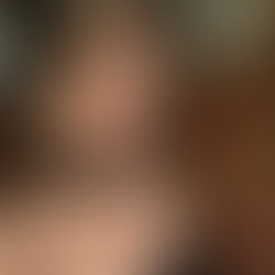 Woman, Sheridan Guerrette, with light hair and hoop earrings poses in front of a marble wall, wearing a black top and necklaces, in an elegant indoor setting.