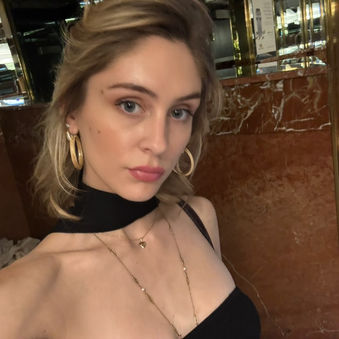 Woman, Sheridan Guerrette, with light hair and hoop earrings poses in front of a marble wall, wearing a black top and necklaces, in an elegant indoor setting.