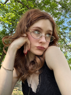 Young woman, Sheridan Guerrette, with glasses rests her chin on her hand under leafy trees, wearing a black lace top and a thoughtful expression.