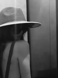 Black-and-white photo of a woman, Sheridan Guerrette, in a wide striped hat and sunglasses, seen from behind near an elevator or doorway.