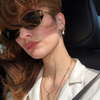 Sheridan Guerrette with red hair in a car, wearing sunglasses and pearl earrings. Wind blows her hair. Dressed in a white satin shirt, relaxed mood.