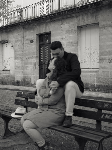 Couple assis sur un banc s'embrassant, chapeau à côté, devant un mur en pierre.
