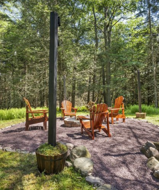 Wooded Poconos fire pit area with Adirondack chairs, gravel circle, and string lights for evening hangouts.