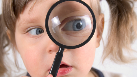 Close-up of a young child holding a magnifying glass up to their eye, enlarging it dramatically while they look curiously at the camera.