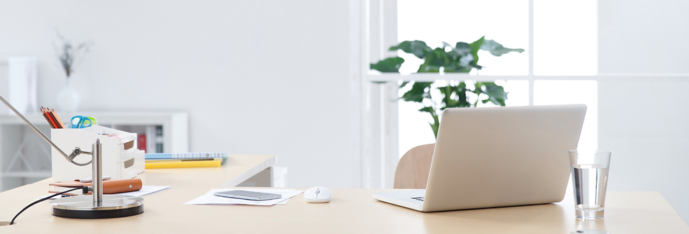 A desk with a laptop, papers, and a glass of water positioned in front of a window with a plant in the background.