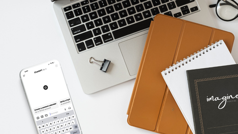 White desk setup with a laptop, an iPhone displaying ChatGPT on it, a tan leather portfolio, and a black notebook with an Imagine Social AI logo on it.