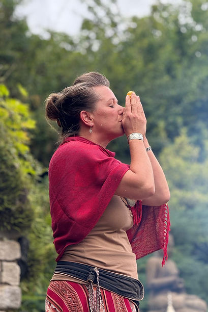 Britta Tanzer in traditionell anmutender Kleidung hält eine spirituelle Zeremonie im Freien ab und hebt die Hände betend vor ihr Gesicht.