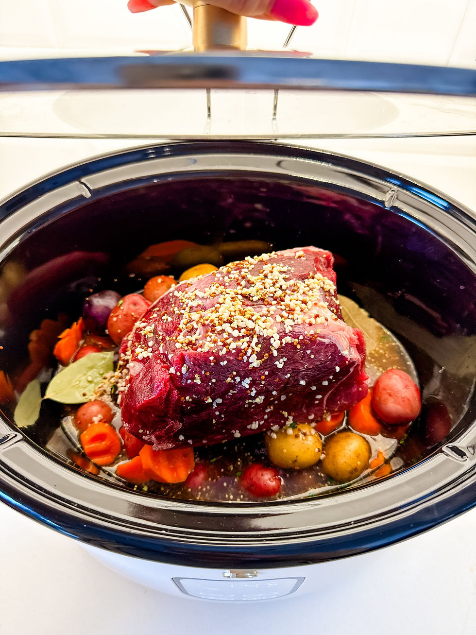 A crockpot holds raw seasoned meat on top of colorful potatoes and carrots. A hand lifts the lid, revealing a kitchen countertop setting.