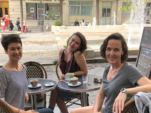 Üç kadın açık hava kafede kahve içiyor; arkada çeşme, sokak ve güneşli şehir meydanı.