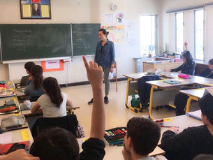 Sınıfta öğretmen kara tahtanın önünde dururken öğrenciler sıralarda ellerini kaldırıyor; güneşli, canlı ders ortamı.