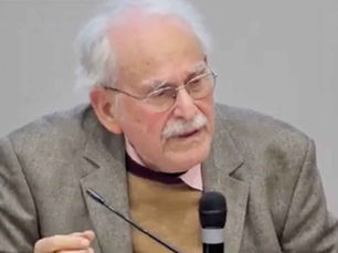 Gri ceketli, gözlüklü yaşlı adam mikrofonda konuşuyor; sade arka planda ciddi bir ifade var. Alberto Eiguer.