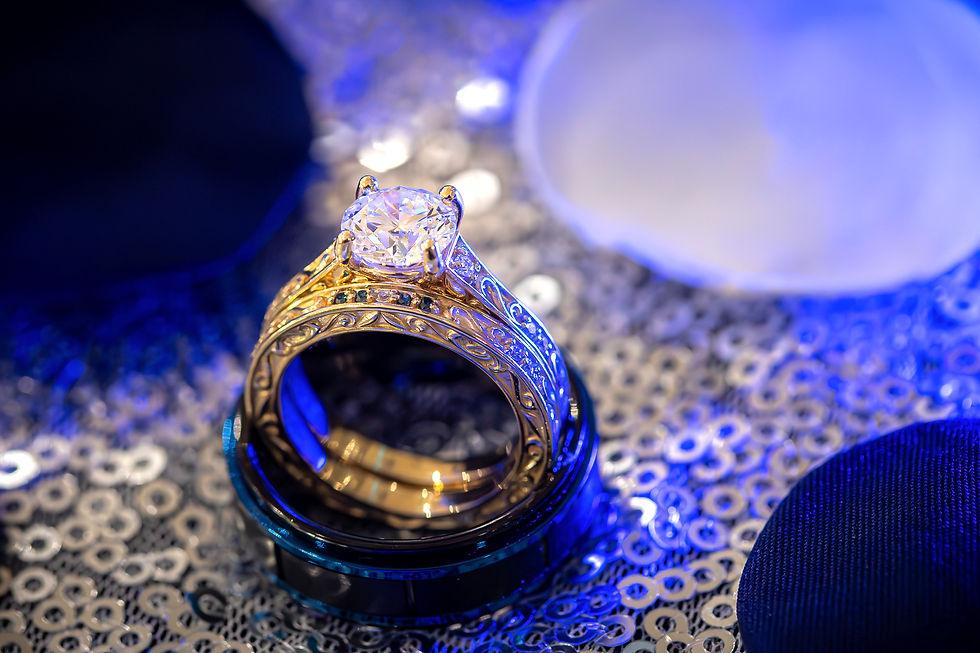 Gold ring with elaborate design and large diamond, set against a sparkling sequin background. Blue lighting creates an elegant ambiance.