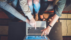Three women lean over a laptop on a coffee table, pointing at the keyboard and screen, in a casual indoor meeting on Bookdio.