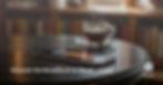 A photograph of a sleek, modern smartphone resting on a dark wooden table. The phone's screen glows with a soft, inviting light, displaying an abstract visualizer representing audio content. A steaming cup of coffee sits slightly behind the phone, casting a warm reflection on the table's polished surface. The background fades into a softly blurred view of a cozy, sunlit study with books lining the walls.