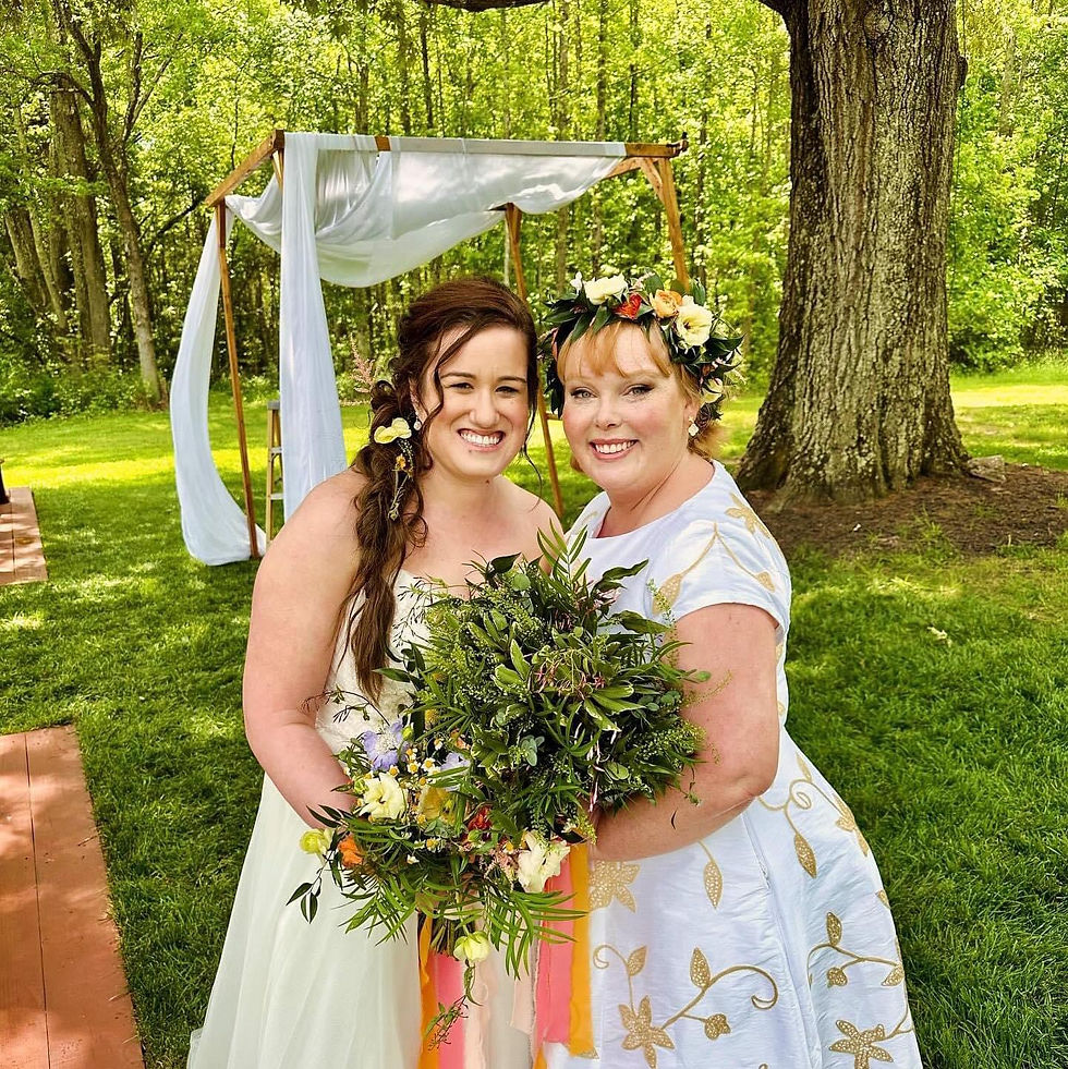 Two brides stand outdoors under a large tree beside a wooden wedding arch draped with white fabric. One wears a strapless white gown with loose waves and side braid, the other wears a white dress with gold floral embroidery and a fresh flower crown. Both hold a large bouquet of greenery and colorful flowers, smiling in the sunlight.