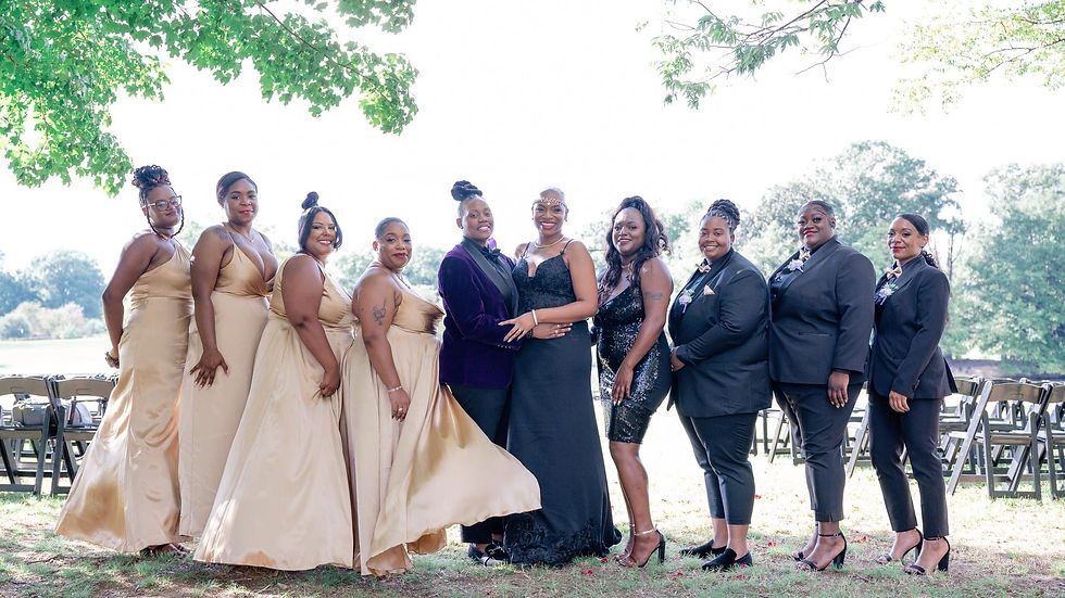A wedding party stands outdoors under a tree, smiling at the camera. The couple at the center wears a dark velvet suit jacket with black trousers and a sleeveless black gown with lace details. Bridesmaids in gold satin dresses stand on one side, while attendants in black suits with floral boutonnieres stand on the other. Folding chairs and trees are visible in the background.