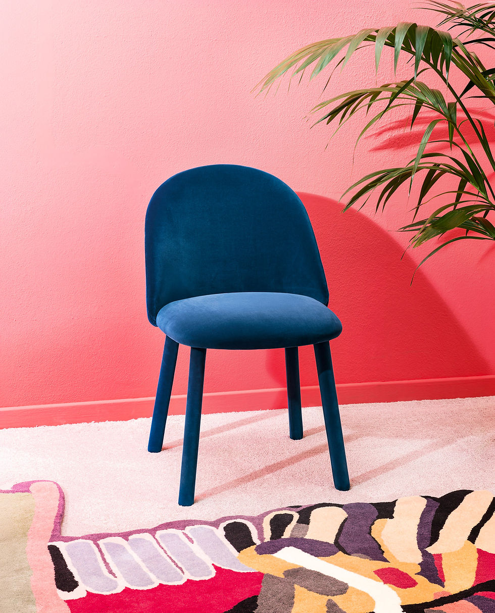 A design furniture photography studio set showcasing a vibrant blue velvet dining chair by Miniforms, positioned over an artistic graphic design rug by Nodus Rug designed by Laureline Galliot against a bright pink wall with green palm leaves, set design, photo direction and styling Laura Pozzi Studio.