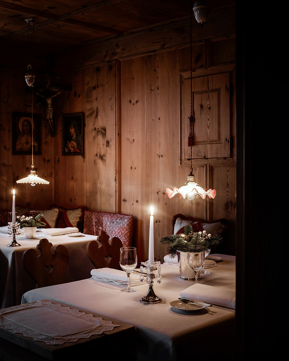 An interior photography close-up focusing on traditionally set restaurant tables with white tablecloths, crystal candelabras, and lit candles inside a historic wood-paneled Stube dining room at Tyrol Hotel in Selva di Val Gardena, photo direction and production Laura Pozzi Studio.