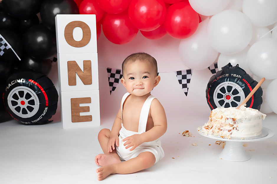 Baby in white romper sits before ONE blocks, balloons and racecar decor, with a smashed cake on a stand.