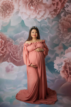 a pregnant woman in a long flowy pink dress standing up against a floral backdrop at her maternity photoshoot in hampshire