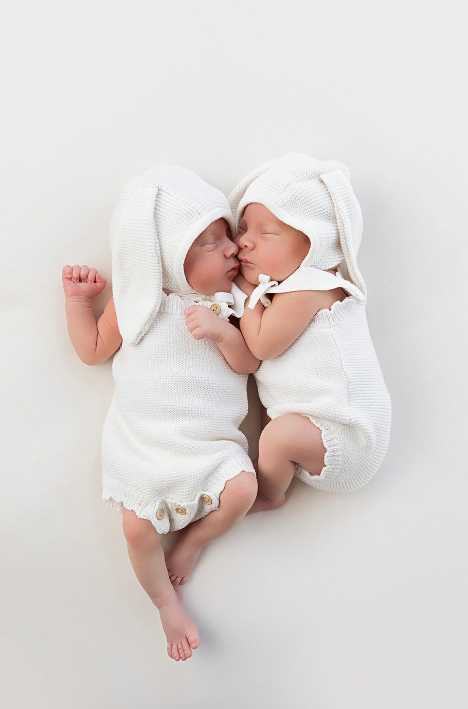 Two sleeping newborn twin babies wearing matching white knitted bunny rompers and floppy bunny hats, posed cuddled closely together on a soft white backdrop during a timeless neutral newborn photography session in Aldershot, Hampshire.