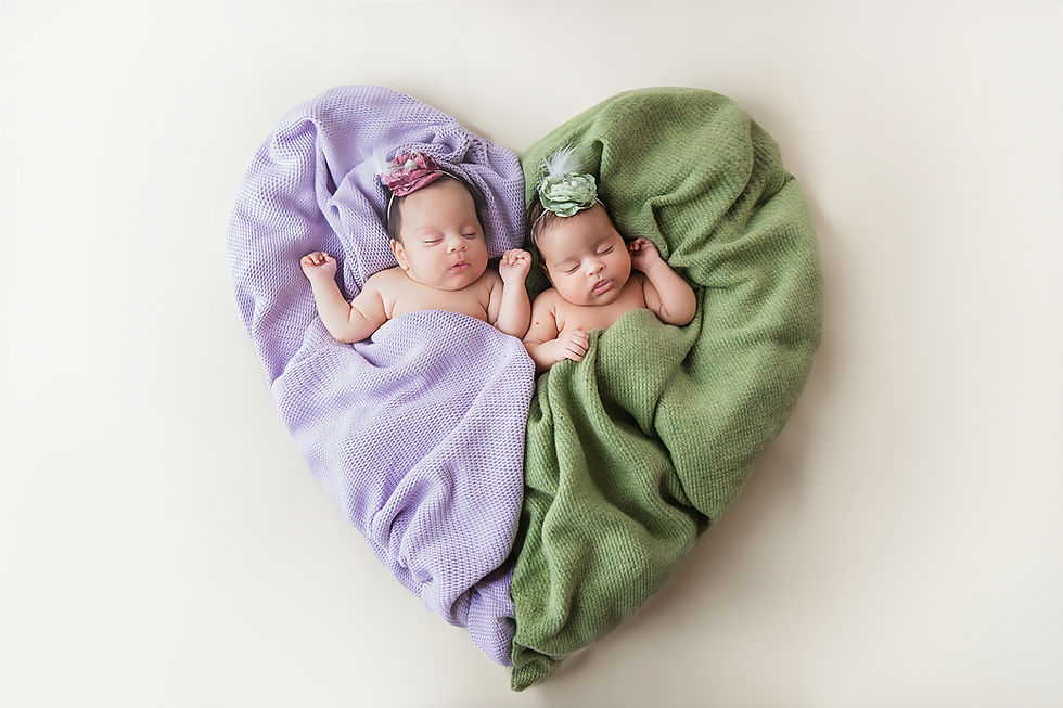 Two sleeping babies, wrapped in purple and green blankets, form a heart shape. Each wears a matching floral headband on a soft, cream background.