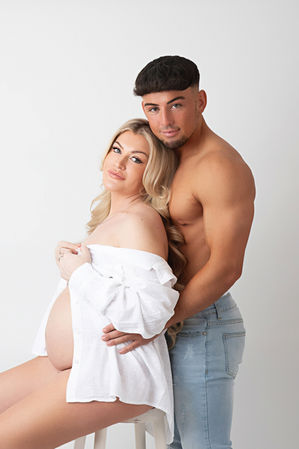 a man and his pregnant wife, both looking at the camera with her sitting on a stool in front of him and them embracing
